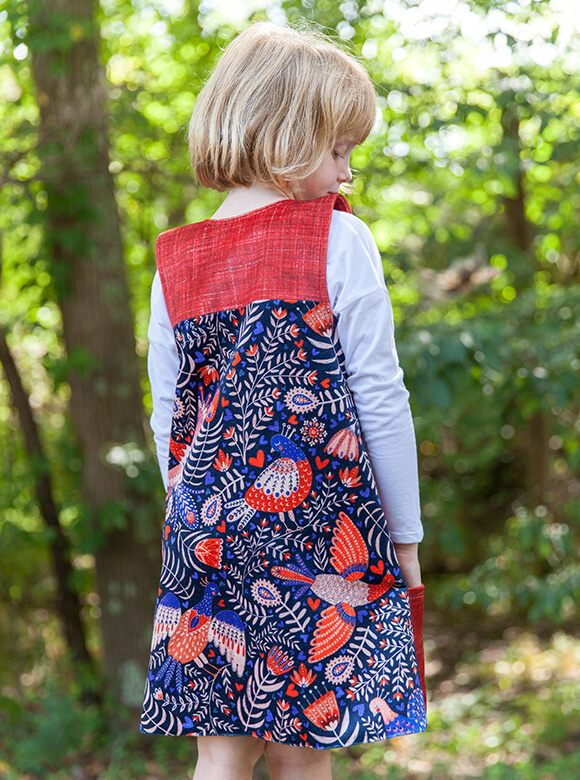Picture of a young, blond girl from behind wearing a romper featuring a blue, white, and red Swedish folk art bird pattern.