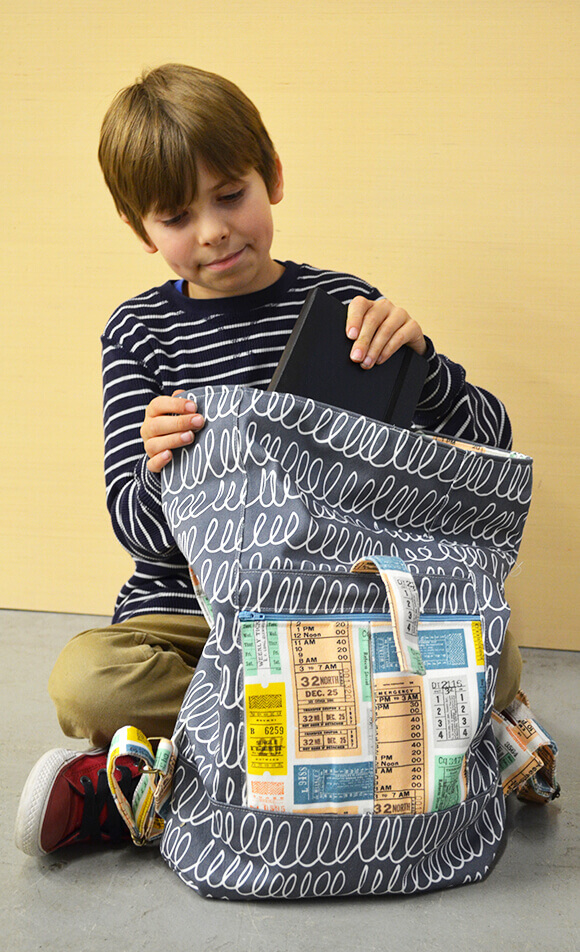 Dogwood Denim Kid’s Backpack Image of a sitting boy putting a book in a black and white scribble patterened kid’s backpack made from Dogwood Denim.