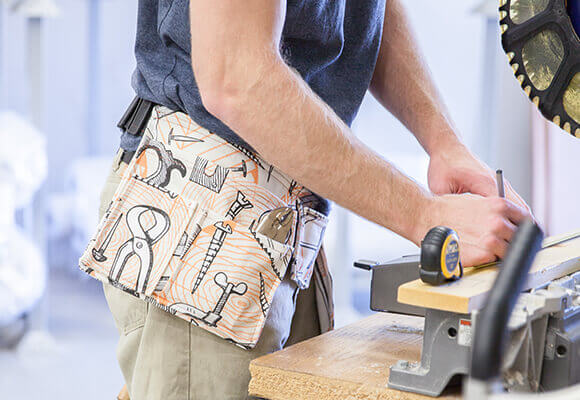 Tool Belt in Dogwood Denim Image of a man measuring a wooden plank, wearing a tool patterned tool belt made from dogwood denim.